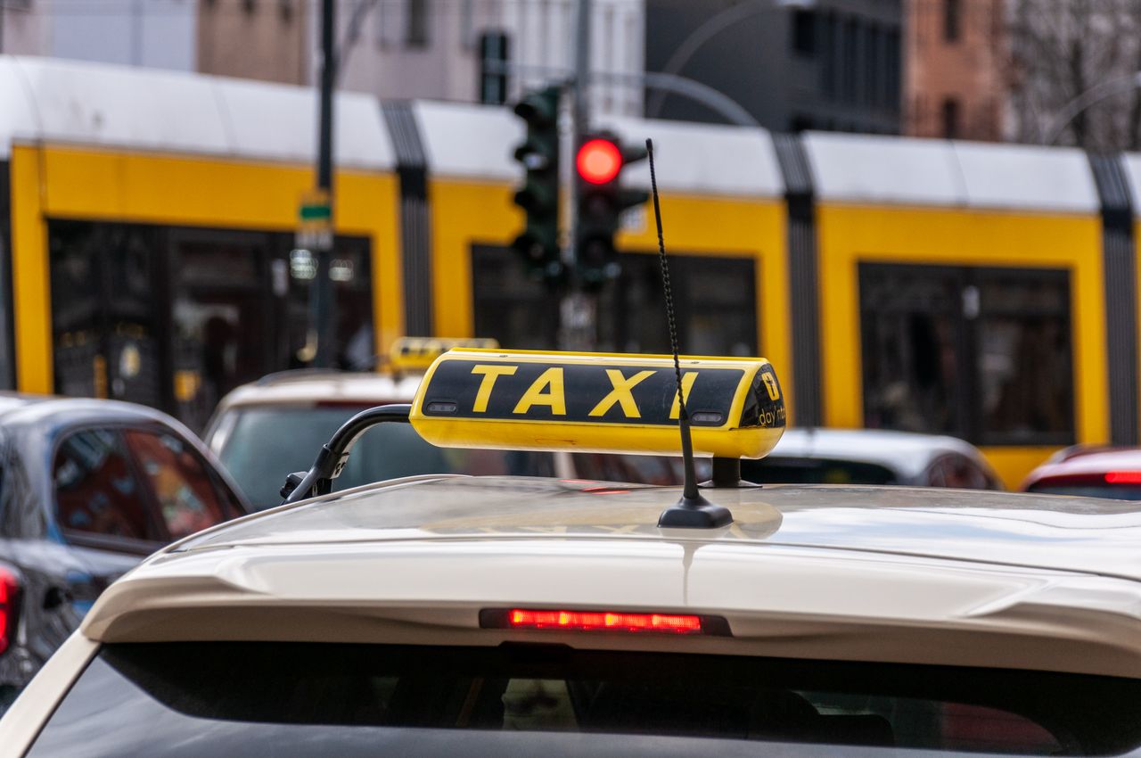 Close-up of a taxi sign with tram and traffic light in city scene.